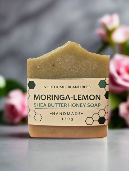 A bar of Moringa-Lemon Shea Butter Honey Soap with a label that includes the brand name and product description, placed on a surface with pink flowers in the background.