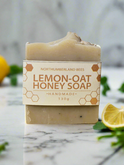 A bar of Lemon-Oat Honey Soap with a label that reads 'Northumberland Bees' and 'Lemon-Oat Honey Soap - Handmade 130g'. The soap is on a marble surface with lemon slices and green leaves in the background.